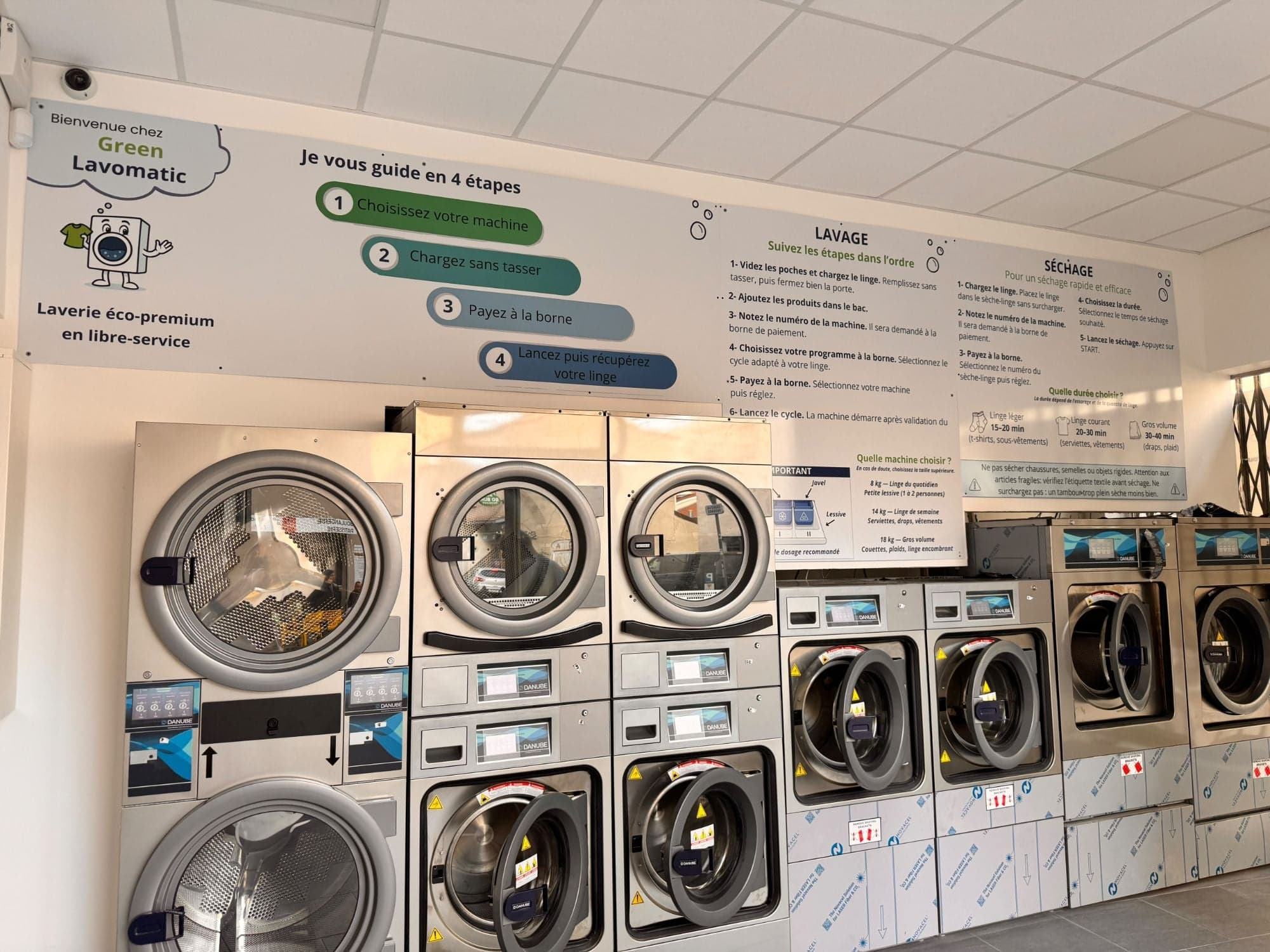 Intérieur de la laverie Green Lavomatic : sèche-linge superposés, machines à laver et panneaux d’explication au mur